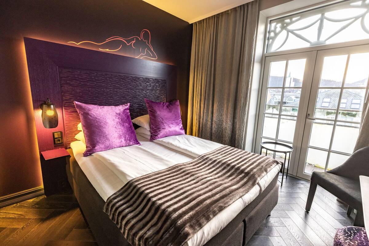 Una habitación de hotel moderna con una cama grande, almohadas moradas, un cabecero con luz de neón, una mesita pequeña y vistas a la ciudad a través de amplias ventanas.