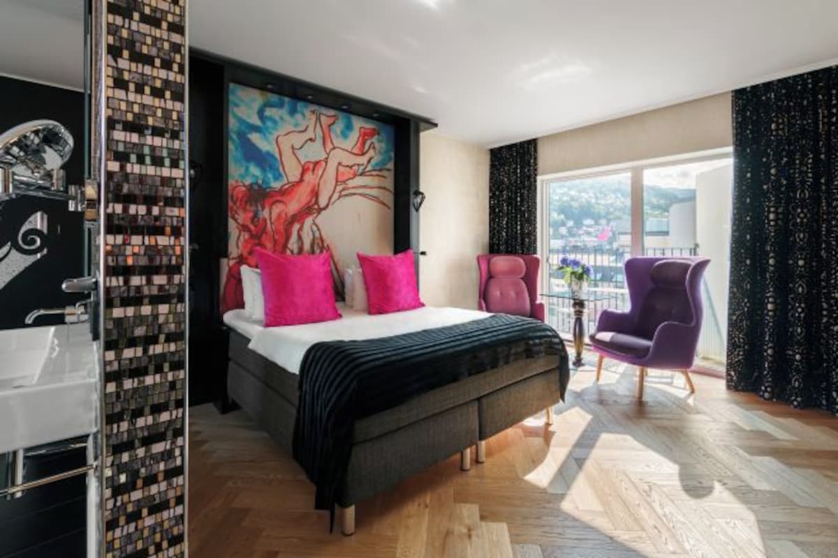 Una habitación de hotel moderna con una cama grande, un baño con espejo y lavamanos, un balcón con vistas, y un mural colorido en la pared.