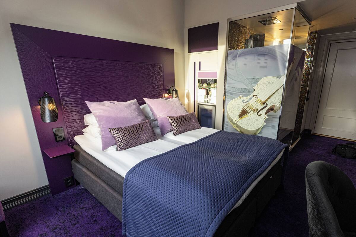 Un dormitorio moderno con cabecera morada, una cama grande con ropa de cama blanca y morada, una manta azul y un box de ducha de vidrio.