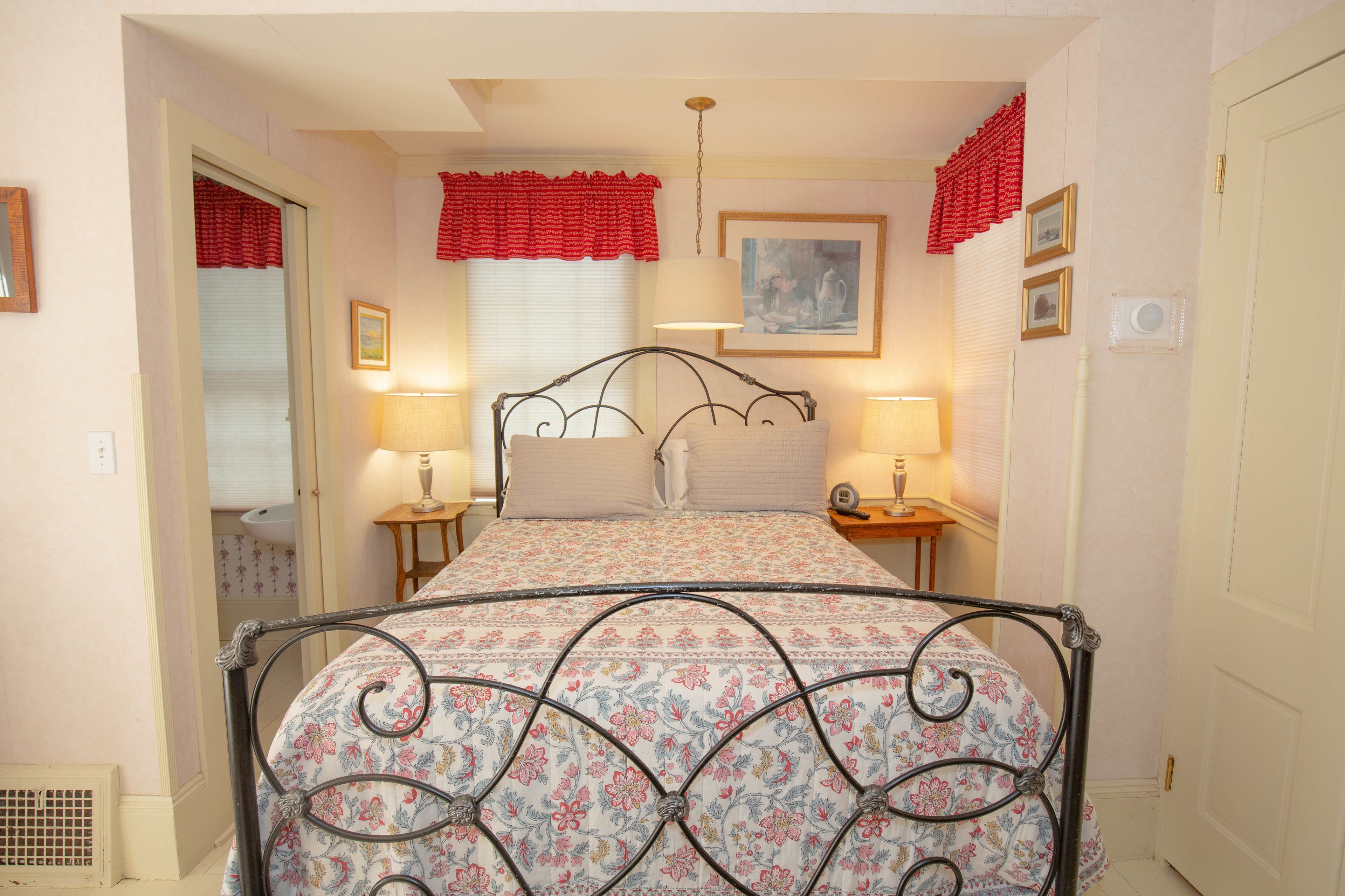 Quarto com colcha florida, estrutura de cama em metal, duas luminárias de cabeceira, janela com cortina vermelha e um quadro na parede.
