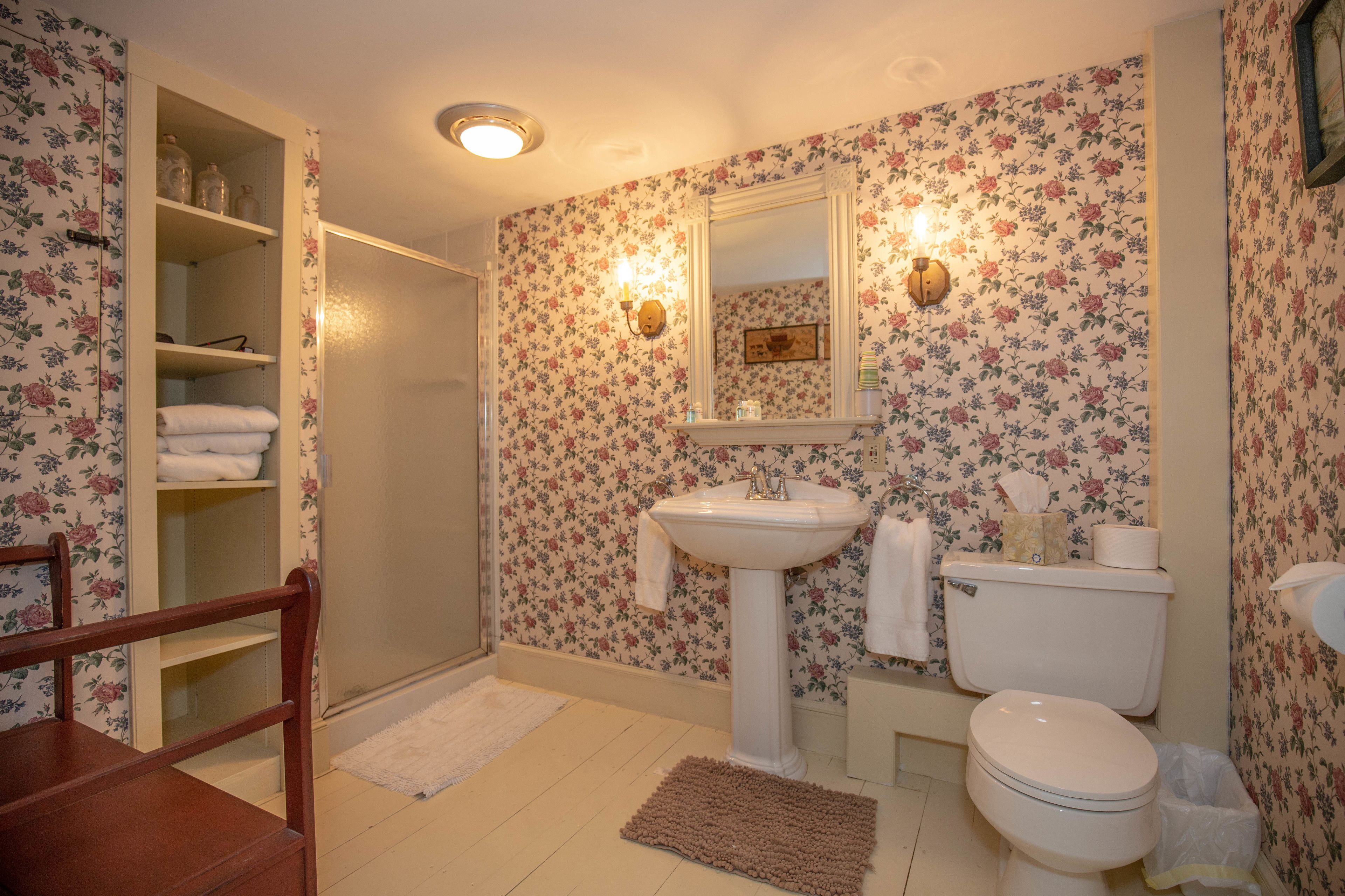 Un bagno con carta da parati a motivi floreali, un lavabo su piedistallo, un water e una sedia di legno.