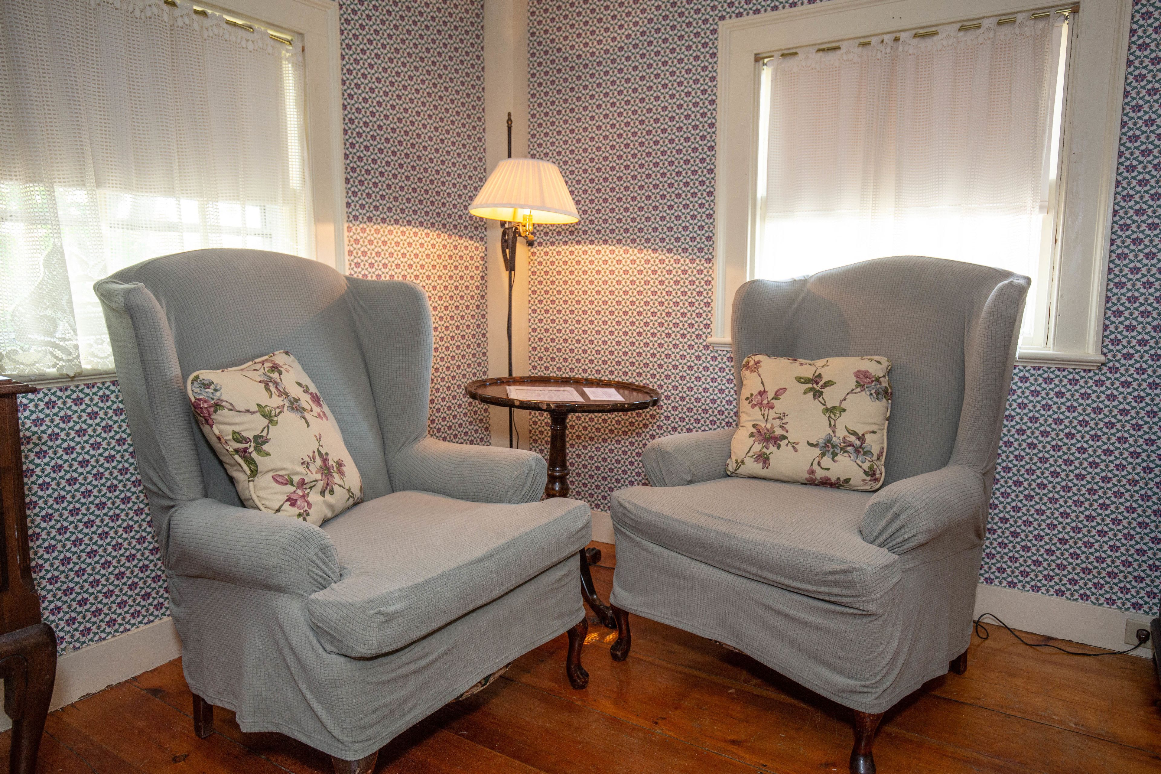 Two upholstered armchairs with floral cushions, a small round table, and a floor lamp in a room with patterned wallpaper and wooden flooring.