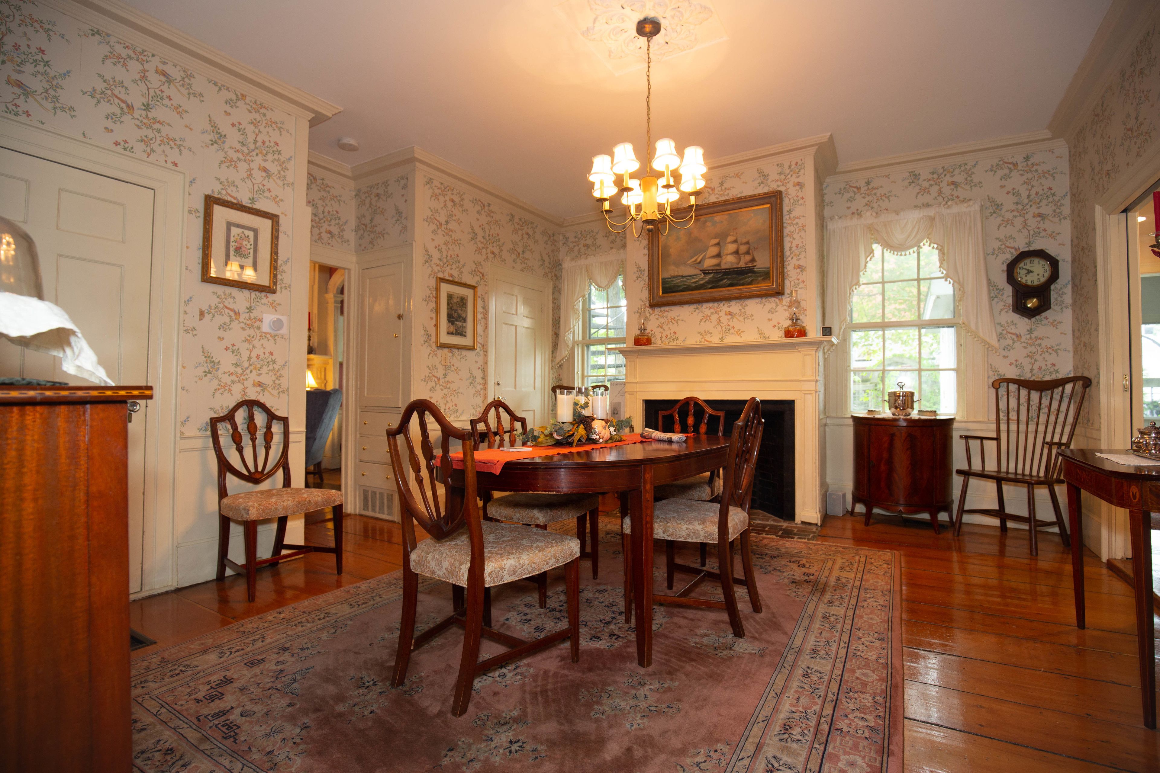 A dining room with a wooden table, chairs, and a chandelier. There is a fireplace with a mantelpiece and a clock on the wall.