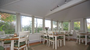 Un espace repas avec des tables et des chaises en bois peintes en blanc, un banc vert rembourré et une vue sur les arbres à travers de grandes fenêtres.