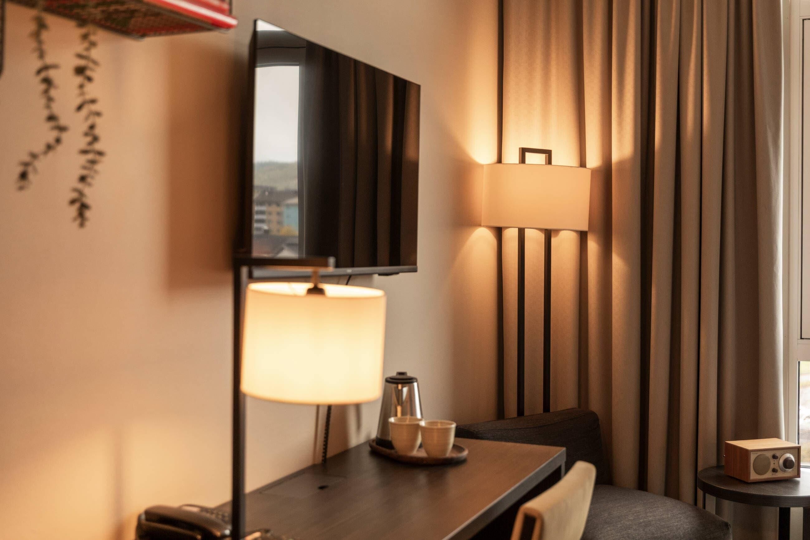 Habitación de hotel con televisor, lámpara, cafetera y una pequeña mesa con tazas.