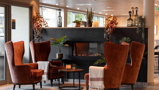 A modern lobby with a fireplace, red velvet chairs, a small table, and decorative plants. - Home Hotel Helma (Mo-I-Rana)
