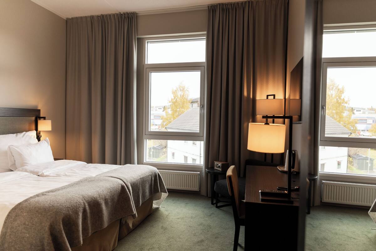 A hotel room with a bed, a desk, a chair, two lamps, and large windows with curtains.