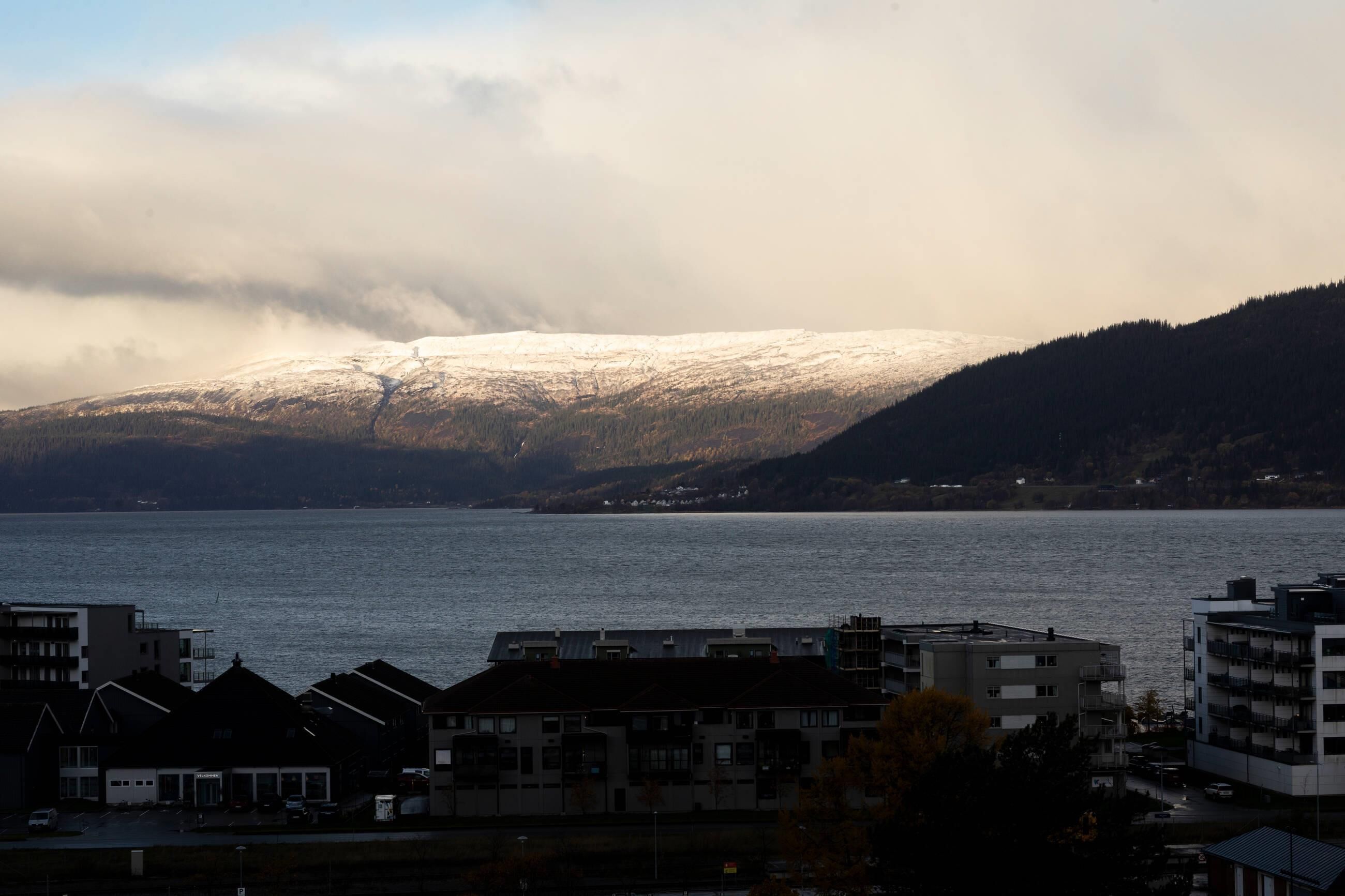 A coastal town with buildings near a body of water, a snow-covered mountain in the background, and a cloudy sky.