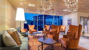 A modern lounge area with sofas, armchairs, and round tables, featuring a large window with a view of buildings. - Home Hotel Helma (Mo-I-Rana)