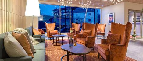 Un moderno espacio de descanso con sofás, sillones y mesas redondas, con un amplio ventanal que ofrece vistas a edificios.
