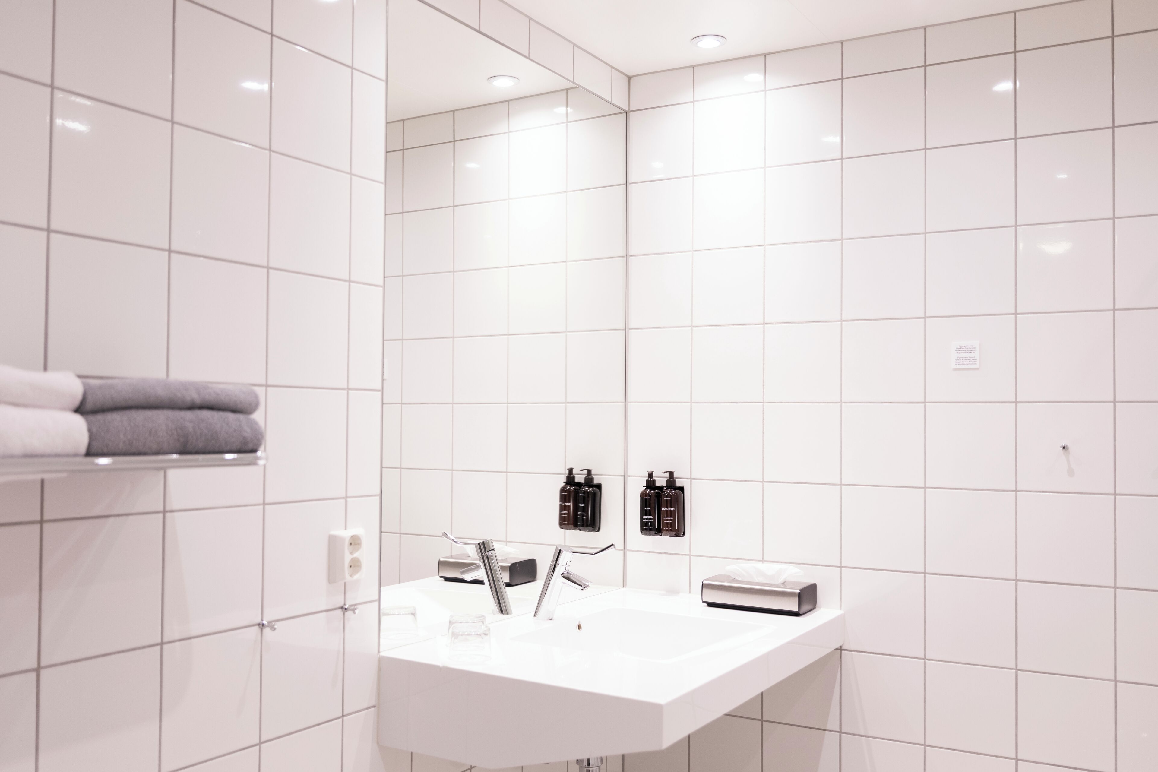Un baño moderno con azulejos blancos, un lavamanos blanco, dos dispensadores de jabón negros y un perchero con toallas dobladas.