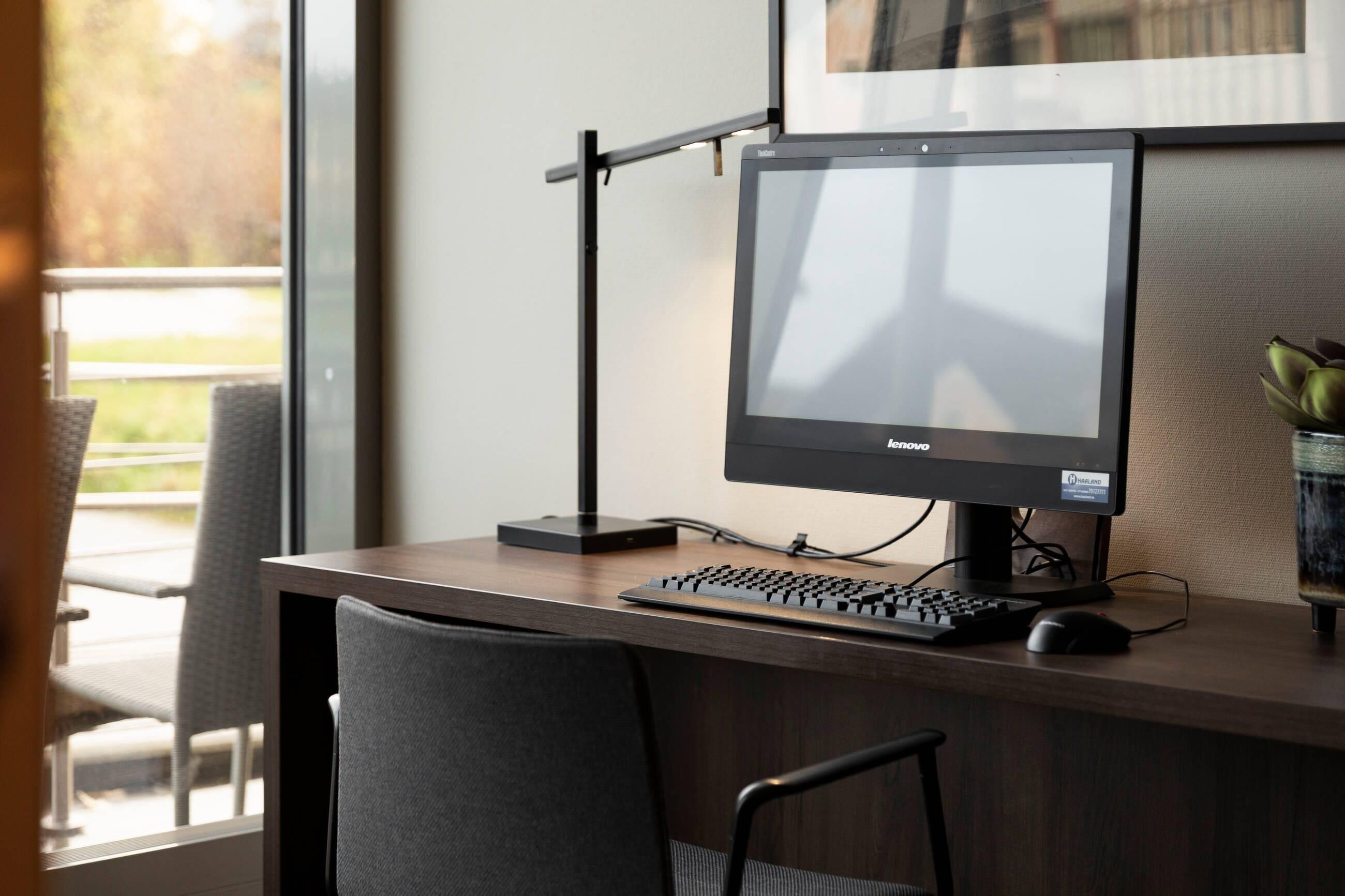 A modern office desk with a Lenovo monitor, keyboard, and mouse, a plant, and a chair.