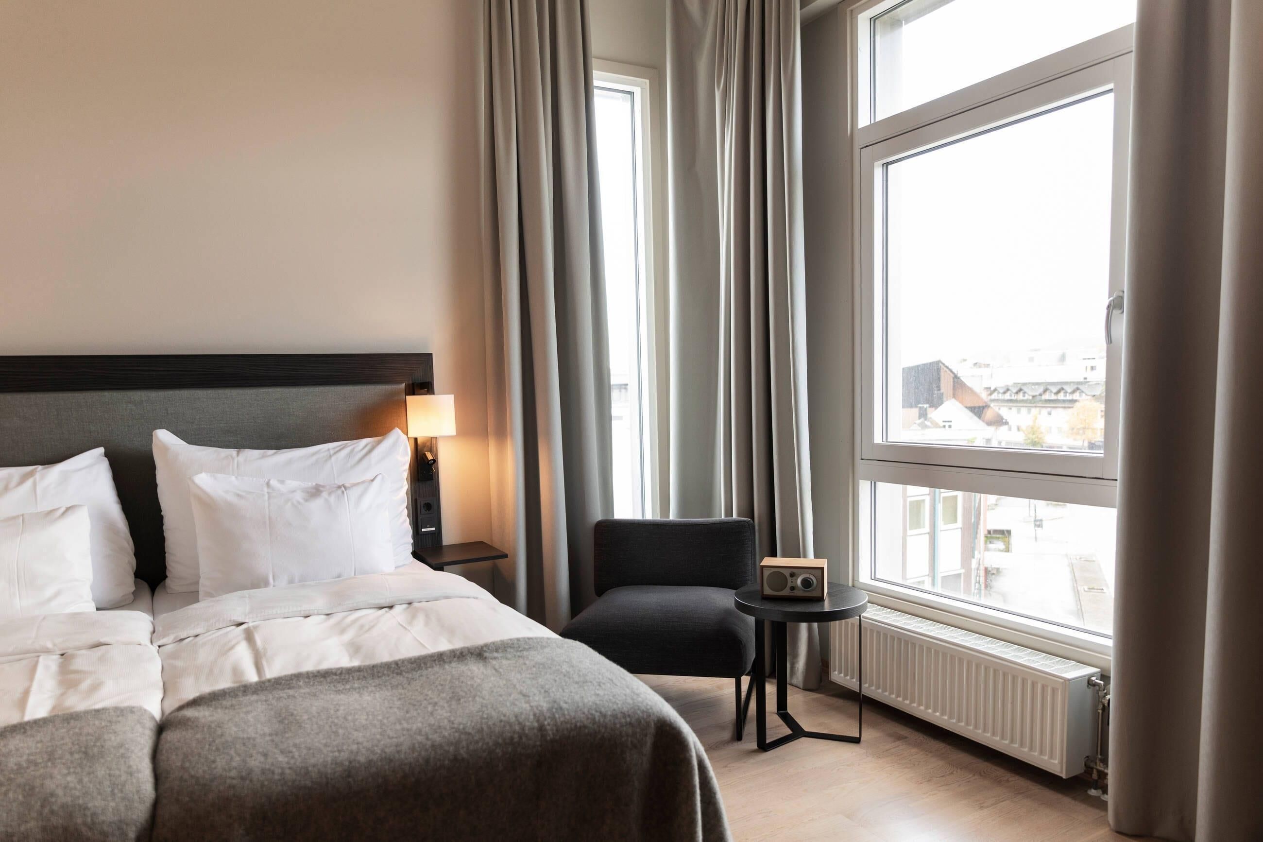 A hotel room with a bed, a chair, a small table with a radio, and a window with curtains.