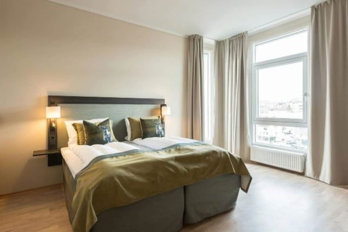 Quarto de hotel com uma cama grande, duas luminárias de cabeceira, vista da cidade através de uma janela ampla e um radiador sob a janela.