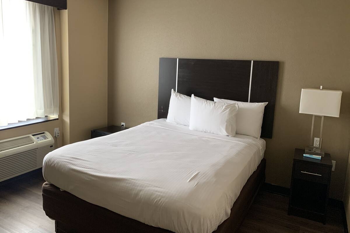 Hotellihuone, jossa on suuri sänky, yöpöytä lampun kanssa, lämmitin ja ikkuna, jossa on verhot.
