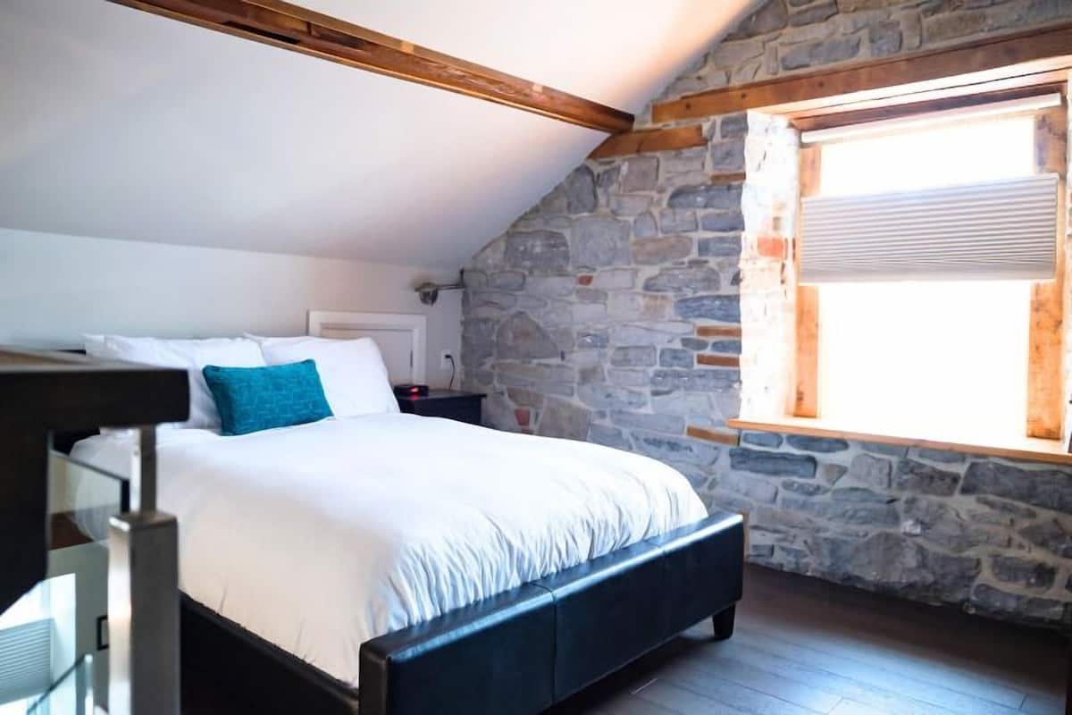 A bedroom with a stone wall, a wooden ceiling beam, a bed with white bedding, and a window with blinds.