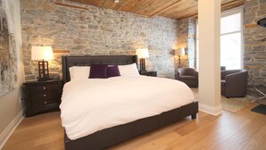 A bedroom with a stone wall, a wooden ceiling, a bed with white bedding, two bedside tables with lamps, and a chair. - Arrow Properties (North Grenville)