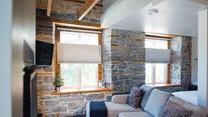 A cozy room with stone walls, a wooden ceiling, a sofa with cushions, a TV, and a window with blinds. - Arrow Properties (North Grenville)