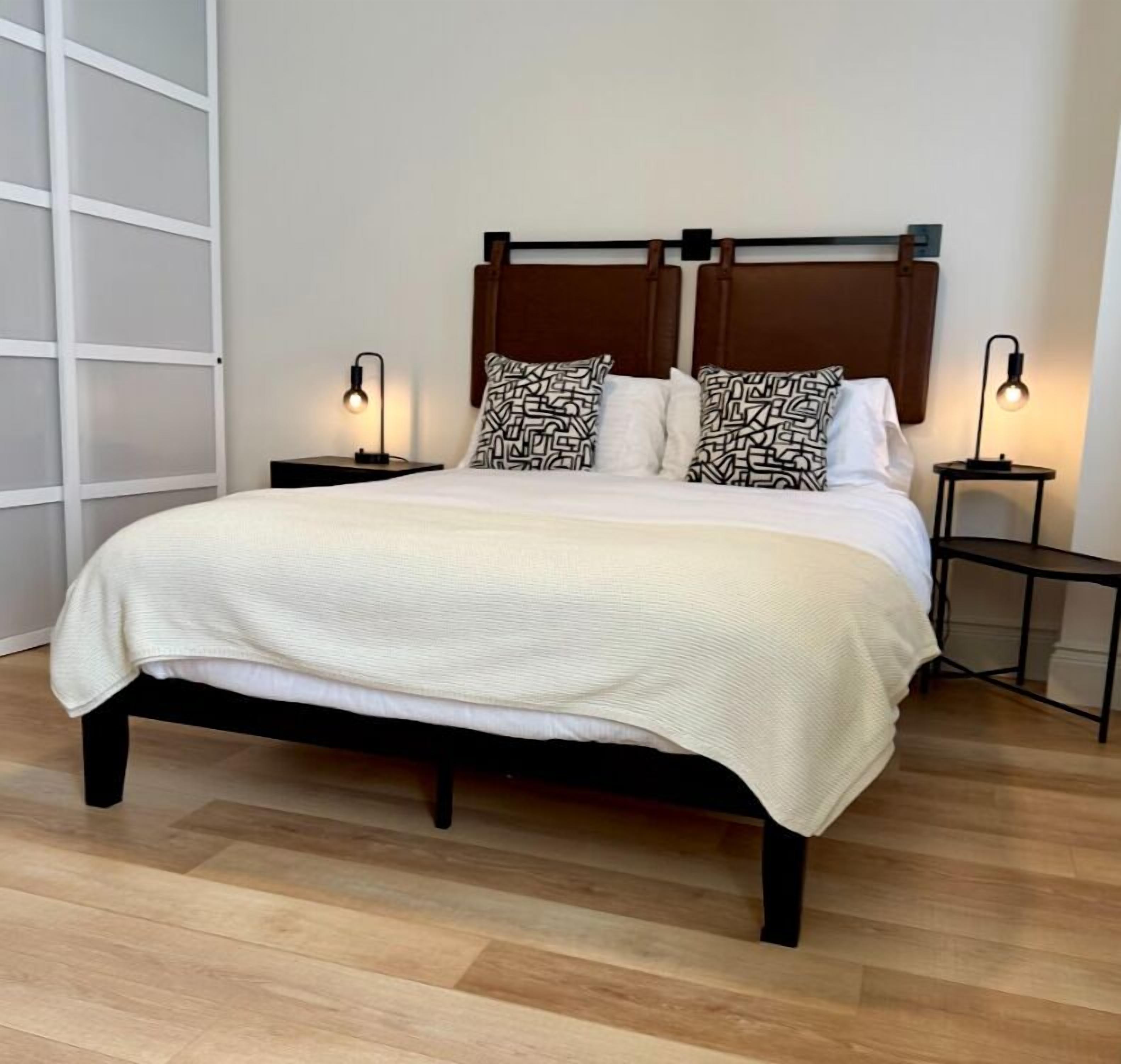 Una cama con estructura de madera, una colcha blanca y almohadas estampadas. Hay dos mesitas de noche con lámparas.