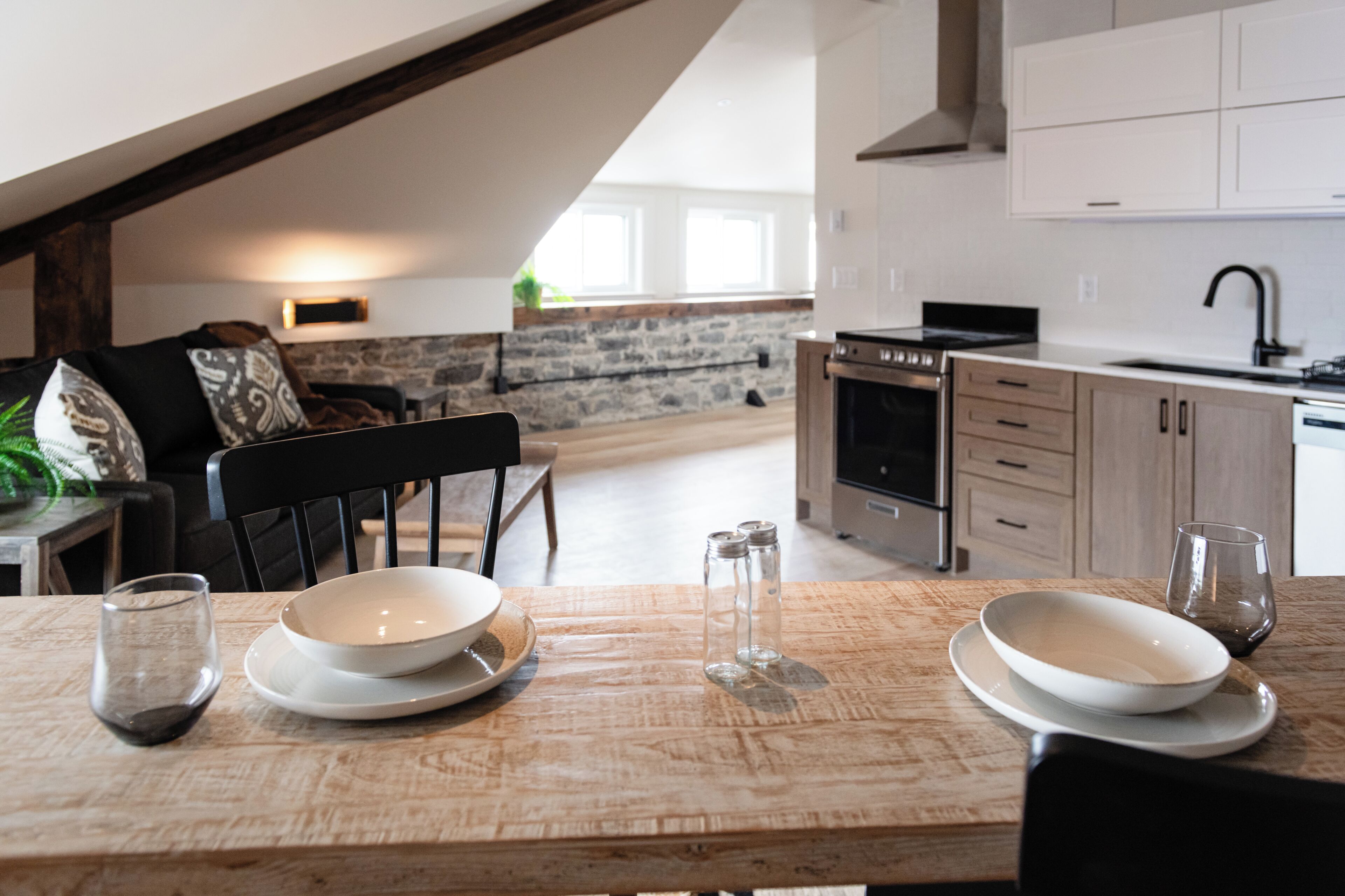 Uma área de refeiç ões com uma mesa de madeira posta para dois, cozinha ao fundo e uma parede de pedra.