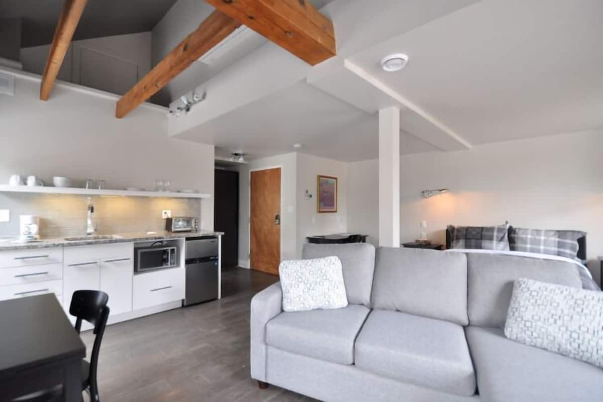 A modern living room with a grey sofa, wooden beams, a kitchen with white cabinets, and a dining area.