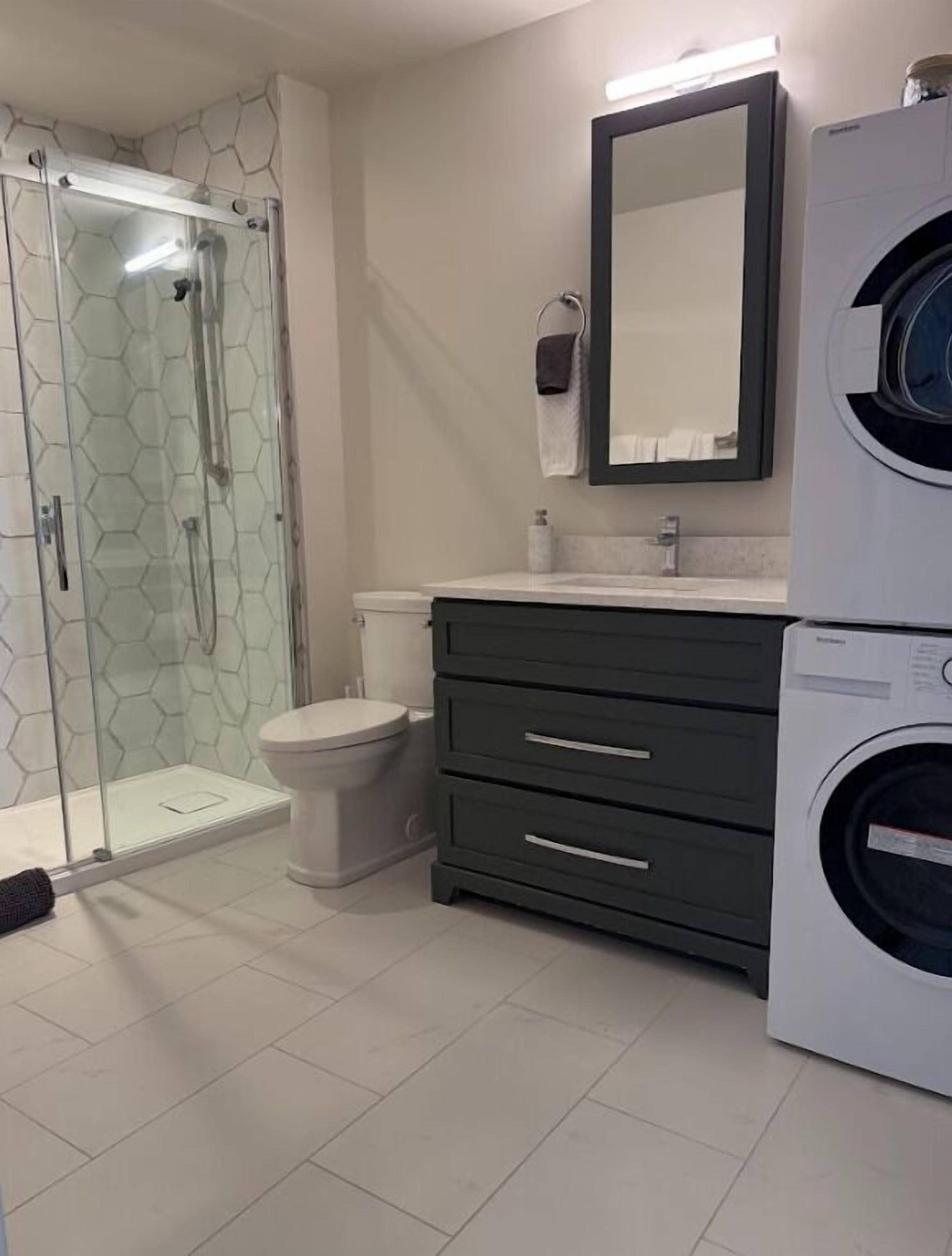 Un baño con cabina de ducha de vidrio, inodoro, lavadora y tocador oscuro con cajones.