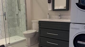 Un baño con cabina de ducha de vidrio, inodoro, lavadora y tocador oscuro con cajones.