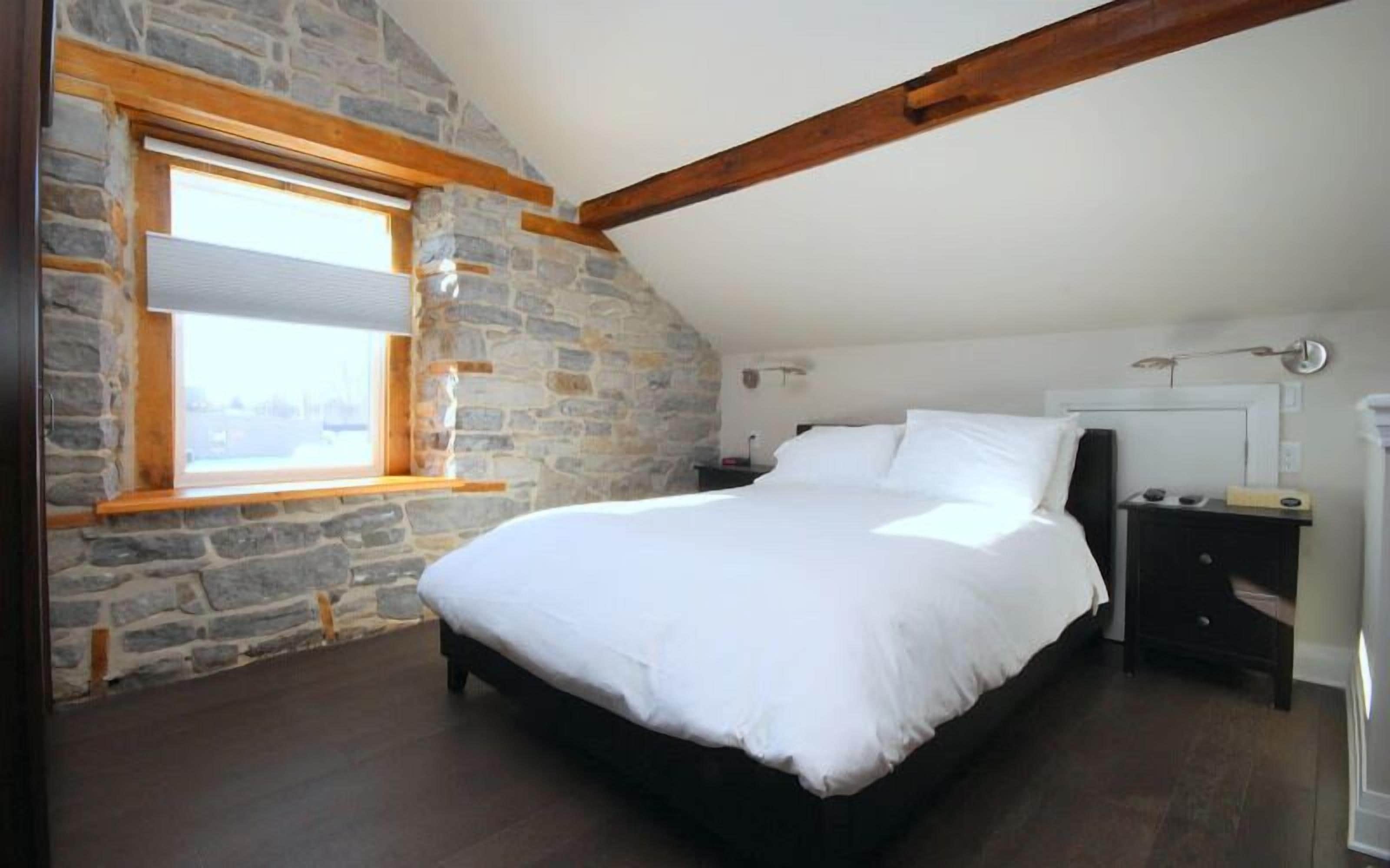 A bedroom with a stone wall, a wooden ceiling beam, a large bed, a nightstand, and a window with blinds.