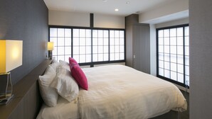 Moderni hotellihuone, jossa on suuri sänky, yöpöytä lampun kanssa ja suuret ikkunat, joista on näkymä.