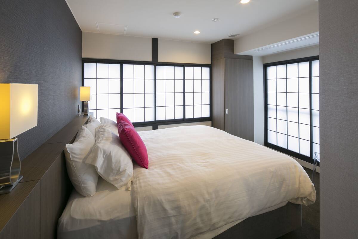 A modern hotel room with a large bed, a bedside table with a lamp, and large windows with a view.