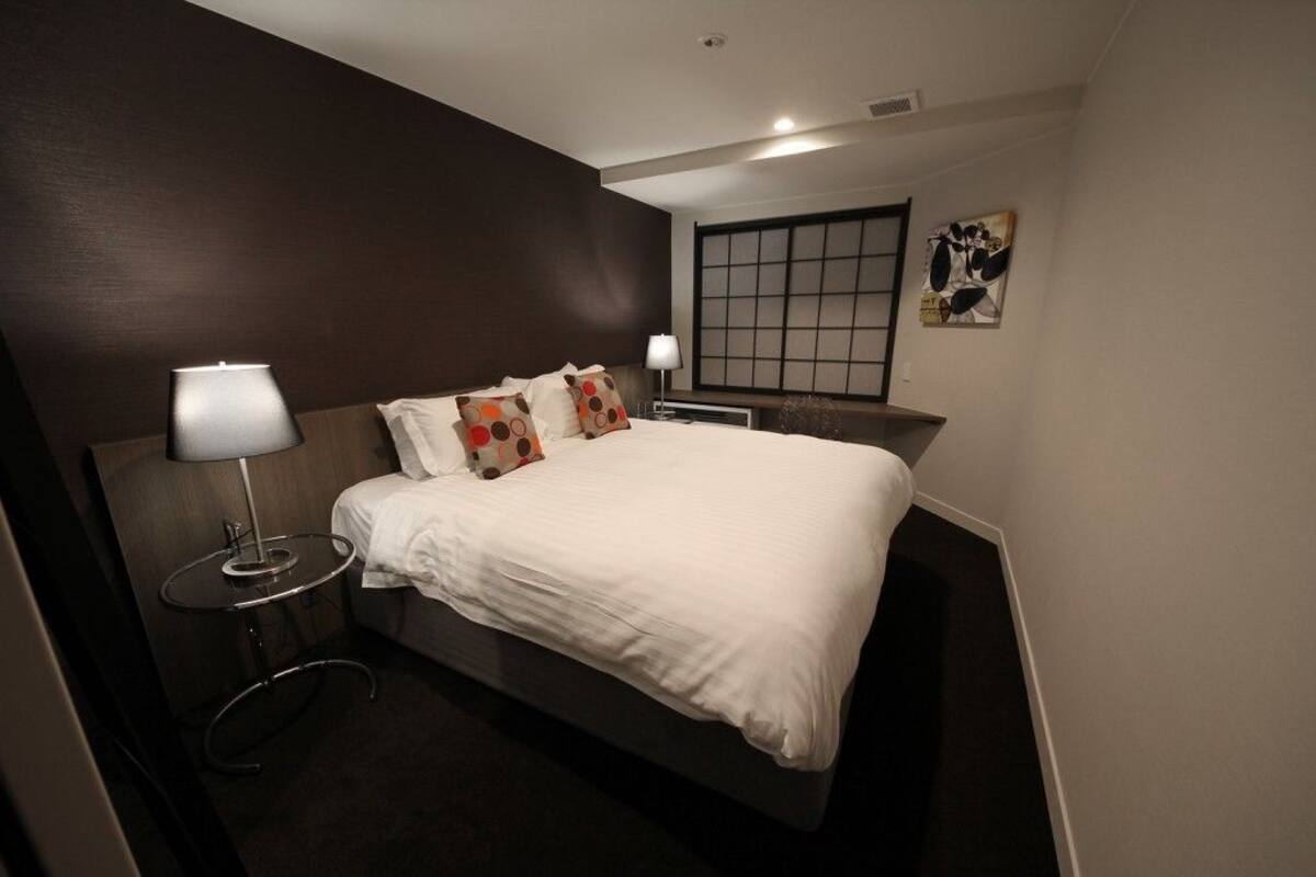 A modern hotel room with a large bed, a bedside table with a lamp, and a view of the city through a window.