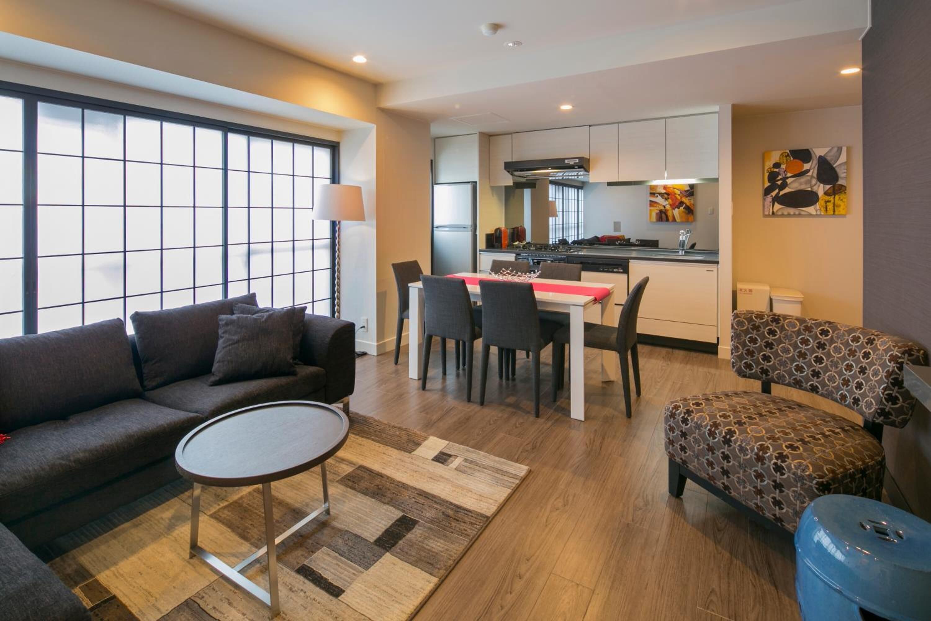 A modern living room with a sofa, a coffee table, and a patterned ottoman. There is a dining area with a table and chairs, and a kitchen in the background.