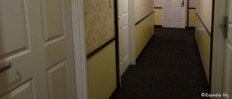 A narrow hotel corridor with doors marked with numbers, carpeted floors, and a ceiling with recessed lighting.