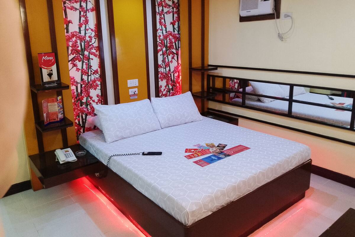 Habitación de hotel con una cama, teléfono en la mesita de noche, una tira de luces roja, un estante con un teléfono y una revista, y un aire acondicionado de pared.