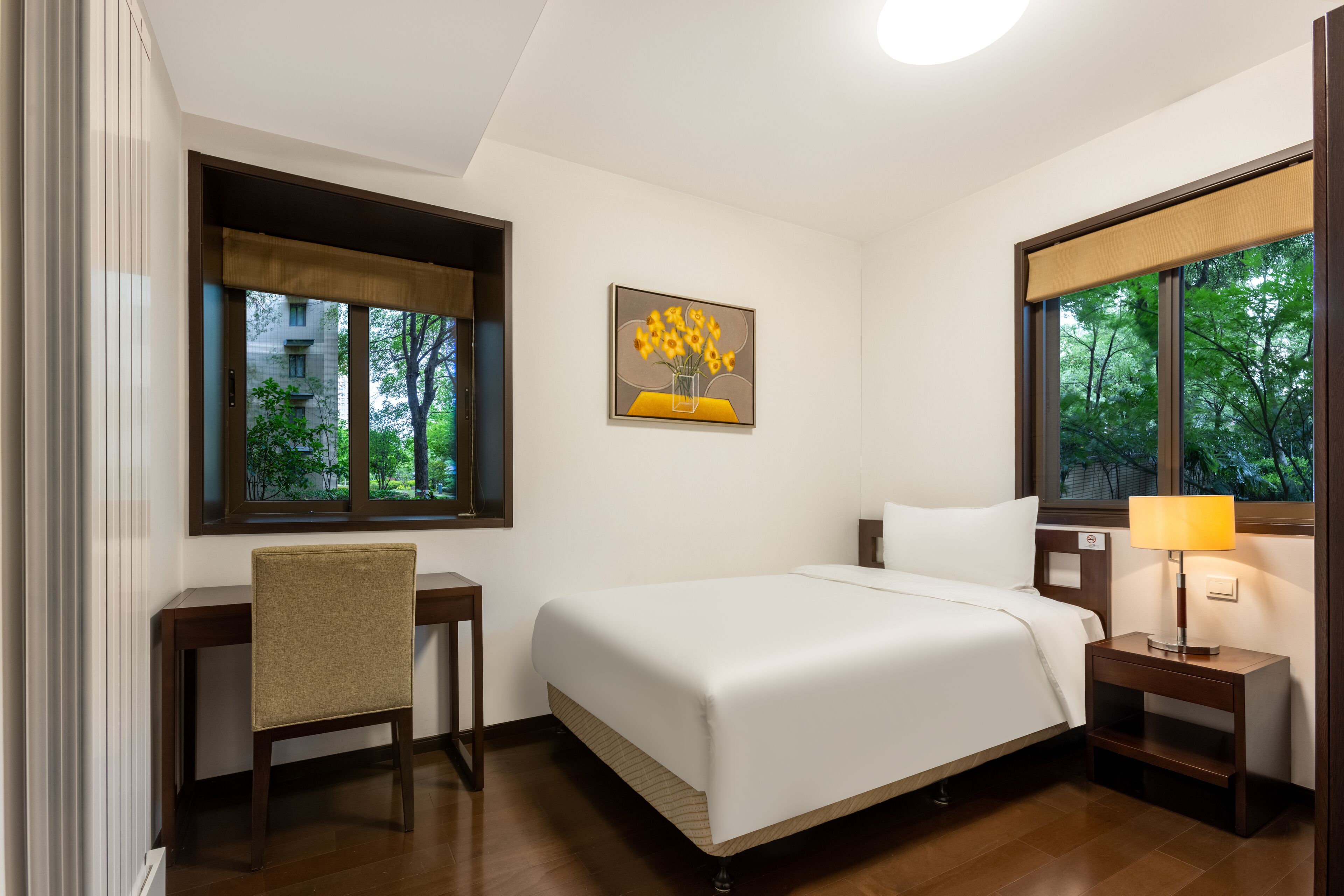 A hotel room with a bed, a desk, a chair, a nightstand with a lamp, and a painting of sunflowers on the wall.