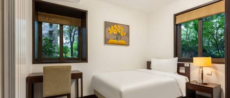 A hotel room with a bed, a desk, a chair, a nightstand with a lamp, and a painting of sunflowers on the wall.