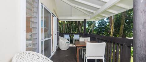 A covered outdoor area with a wooden deck, wicker chairs, a table, and a sliding glass door leading to a room.