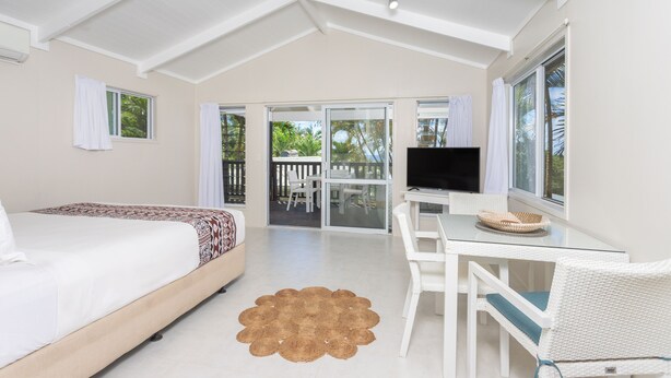 A bedroom with a bed, a TV, a dining table, and a balcony with a view of greenery. - Muri Lagoon View Bungalows (Rarotonga)