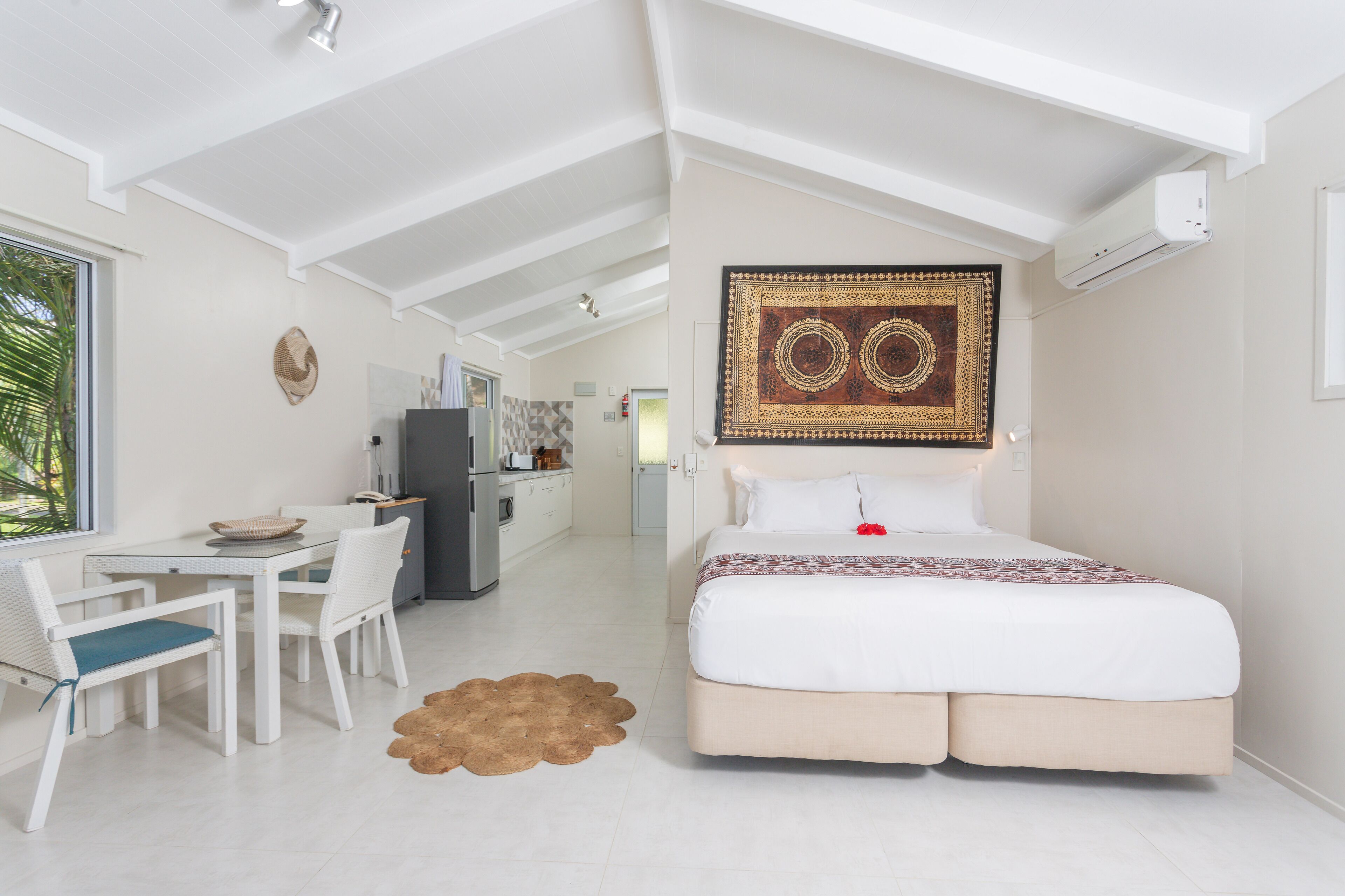 Habitación de hotel moderna con una cama grande, un área de comedor con mesa y sillas, un rincón de cocina y vistas a palmeras.
