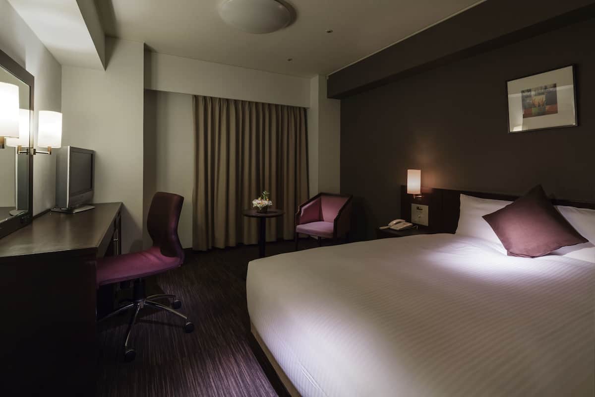 A hotel room with a large bed, a desk with a TV, two chairs, a small table with flowers, and a painting on the wall.