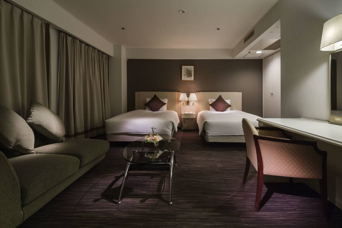A hotel room with two beds, a desk, a chair, a coffee table, and a large window with curtains.