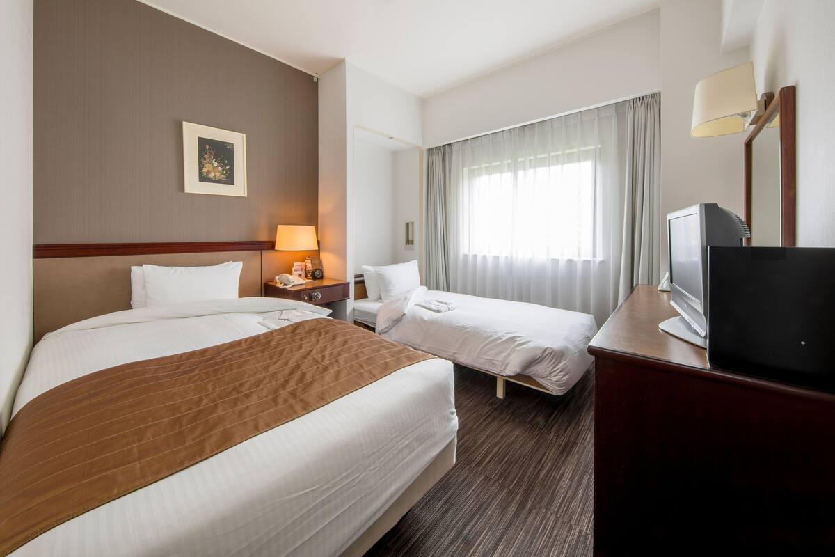 A hotel room with two beds, a television, a desk, and a window with curtains.