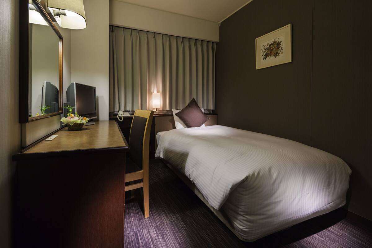 A hotel room with a bed, a desk with a computer, a chair, a lamp, and a window with curtains.