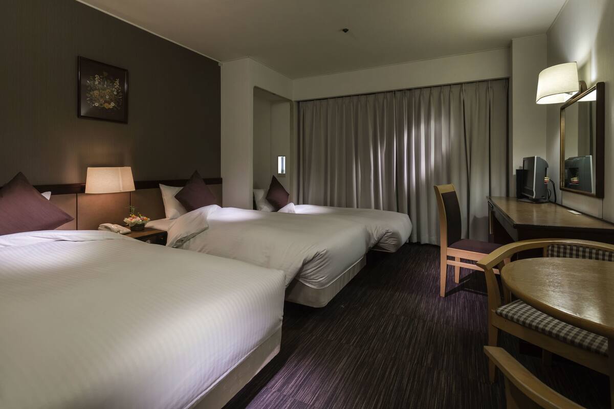 A hotel room with two beds, a desk with a chair, a lamp, a television, and a large window with curtains.