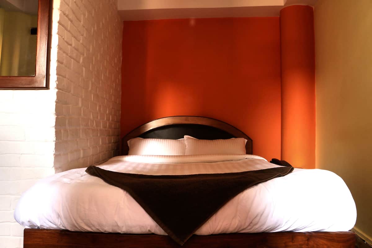 Una cama con ropa de cama blanca y un plaid oscuro, frente a una pared de ladrillo y cortinas anaranjadas.