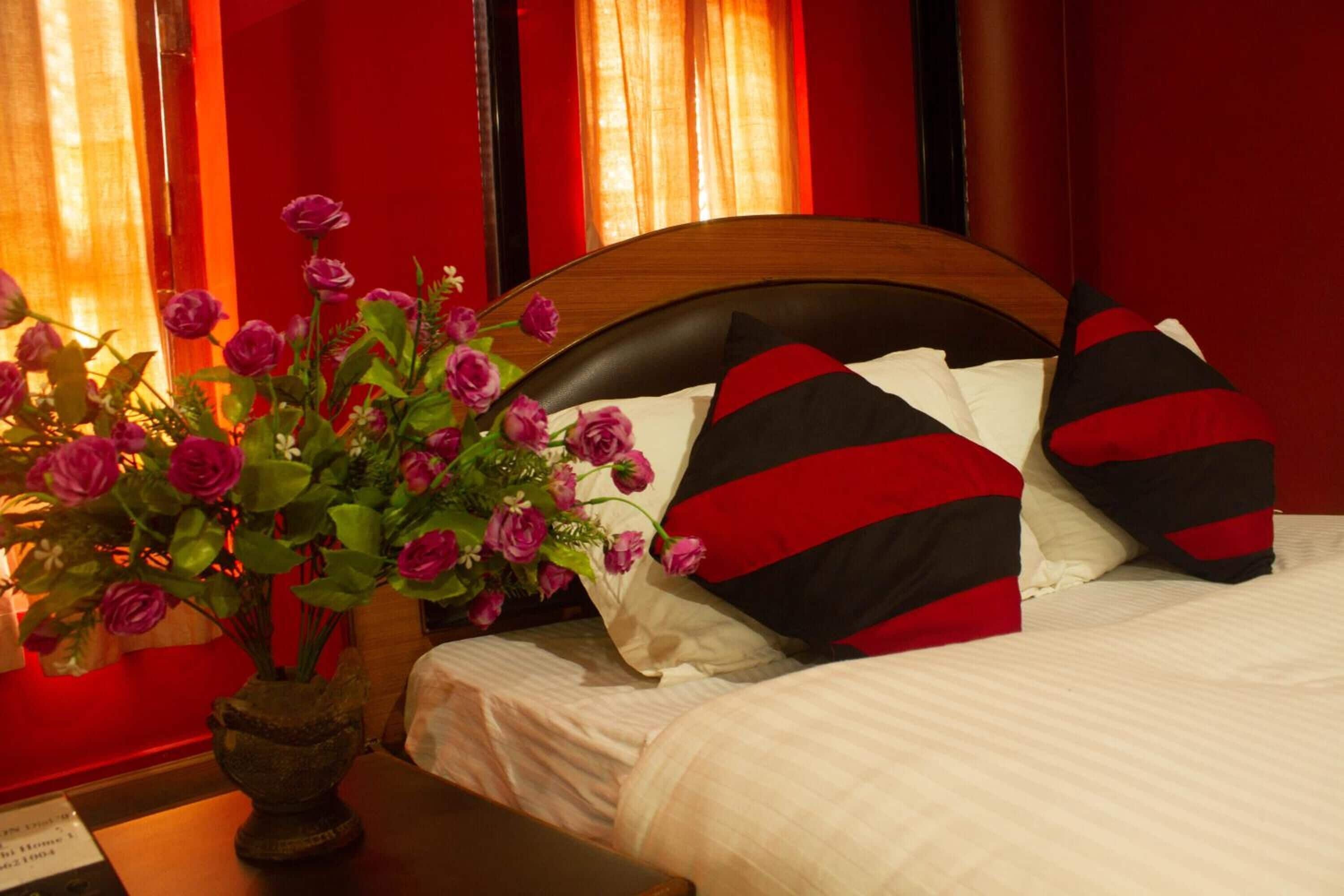 A hotel room with a bed, red curtains, and a vase of purple flowers on a wooden nightstand.