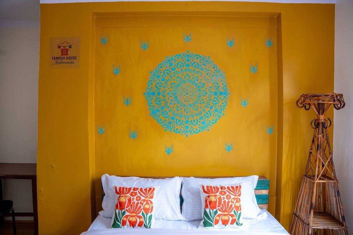 A hotel room with a yellow accent wall, a bed with white bedding and floral pillows, a decorative circular pattern on the wall, and a bamboo stool.