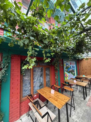 An outdoor seating area with wooden tables and benches, surrounded by greenery and a red brick wall. - Famous House Kathmandu - Hostel (Kathmandu)