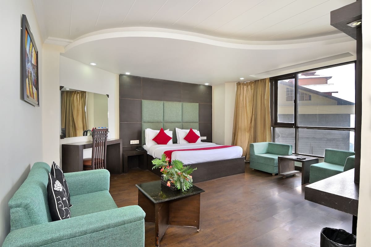 Habitación de hotel con una cama, dos sillones verdes, una mesita con una planta, un escritorio y un ventanal con cortinas.