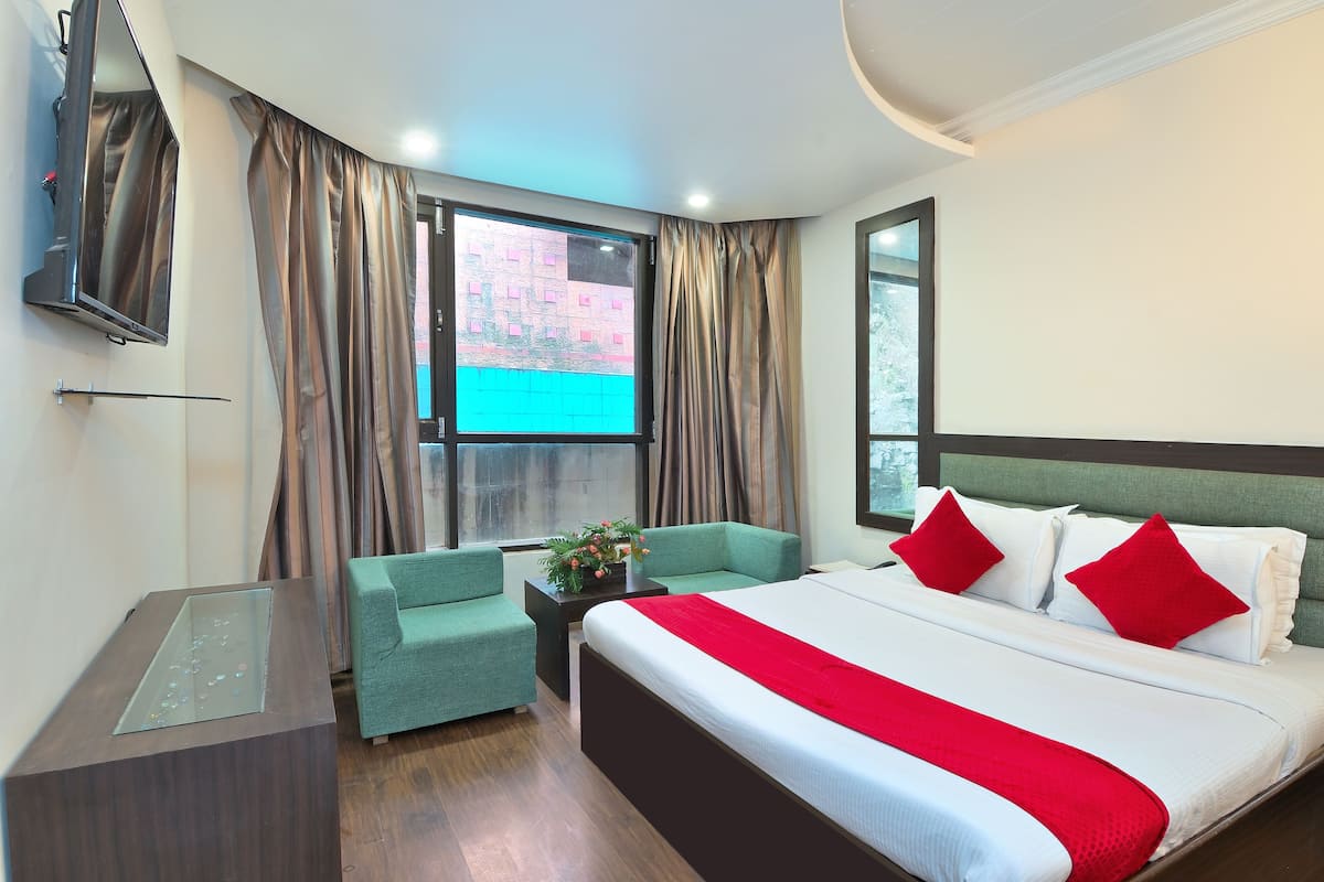 Habitación de hotel con una cama grande, un sillón verde, una mesita con un jarrón y un televisor montado en la pared.
